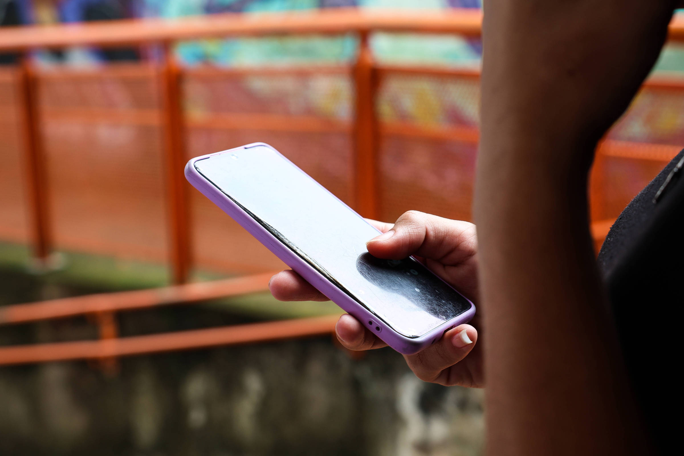 A imagem mostra uma mão segurando um celular com uma capa roxa. O fundo apresenta uma estrutura de metal laranja e uma parede com grafite colorido. A tela do celular está refletindo a luz, tornando difícil ver o que está na tela.