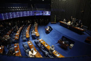 Plenário do Senado Federal com senadores sentados em bancadas curvas de madeira e cadeiras azuis. Painel eletrônico exibe informações legislativas na parede ao fundo. Mesa diretora com cinco pessoas sentadas à direita. Ambiente interno com iluminação artificial e piso azul.