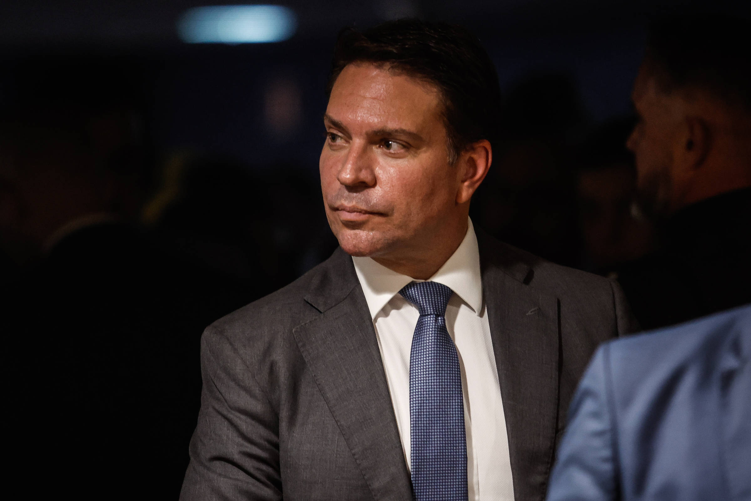 Um homem em traje formal, usando um terno cinza e uma gravata azul, está em um ambiente com pouca luz. Ele tem uma expressão séria e está olhando para o lado, enquanto outras pessoas estão ao fundo, desfocadas. A iluminação suave destaca seu rosto e parte do terno.