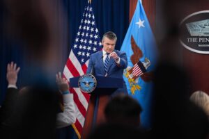 Homem em terno azul escuro e gravata listrada está atrás de um púlpito com o selo do Departamento de Defesa dos EUA. Ele aponta para alguém na plateia durante coletiva, com as bandeiras dos Estados Unidos e da Força Aérea ao fundo.