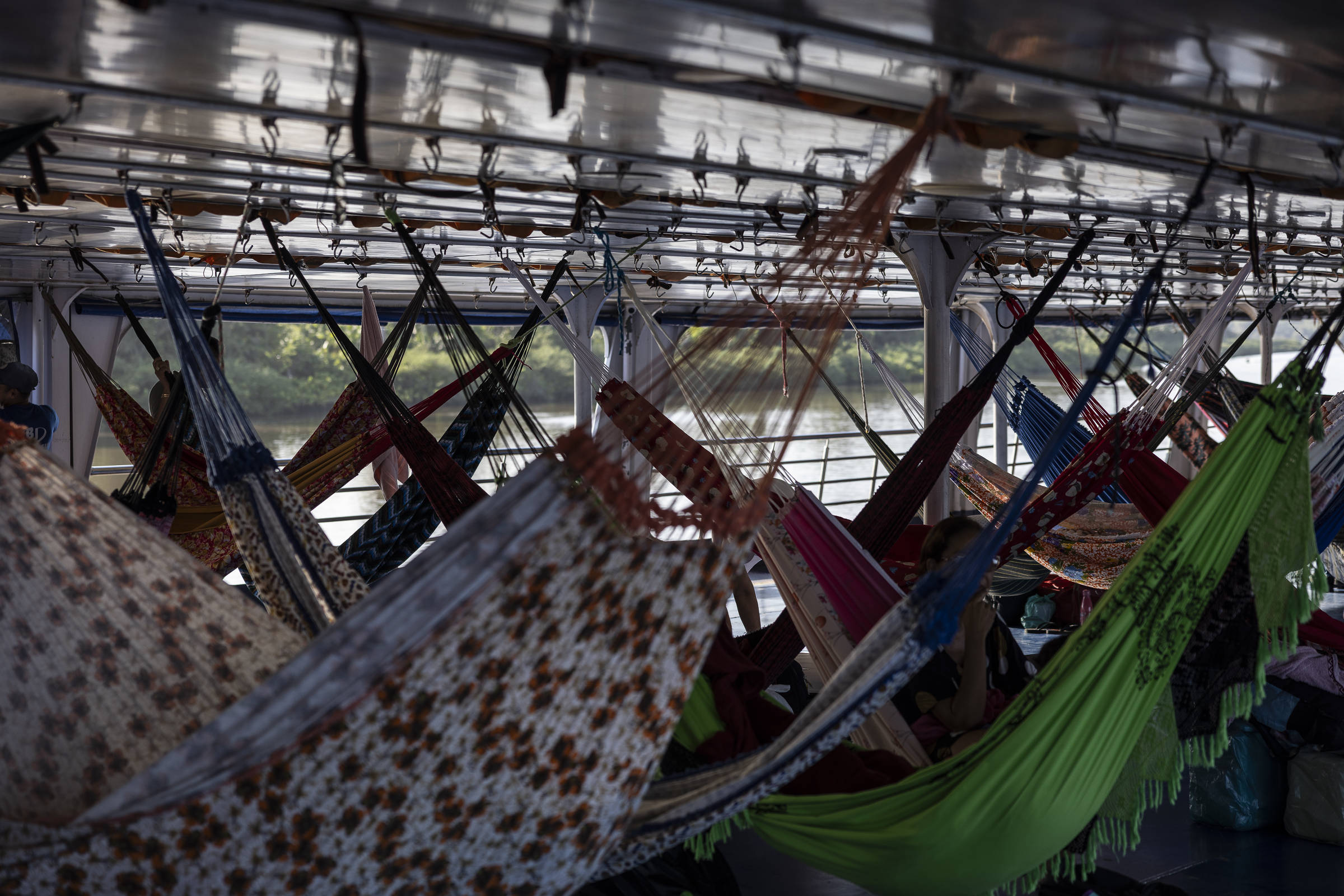A imagem mostra o interior de um barco, onde várias redes estão penduradas. As redes são de diferentes cores e padrões, incluindo flores e tons sólidos. Ao fundo, é possível ver janelas que dão para o exterior, onde há vegetação e água visíveis. A iluminação é suave, sugerindo um ambiente tranquilo.