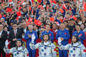 Três astronautas chineses em trajes espaciais acenam para uma multidão. Ao fundo, pessoas vestindo uniformes azuis e civis seguram pequenas bandeiras da China, celebrando o evento.
