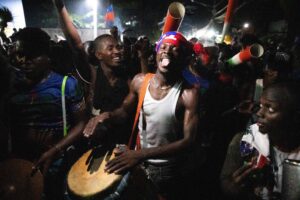 Homens em festa noturna tocam tambores e instrumentos de percussão em ambiente ao ar livre. Um homem no centro usa faixa vermelha na cabeça e camiseta regata branca, expressando energia enquanto toca o tambor.