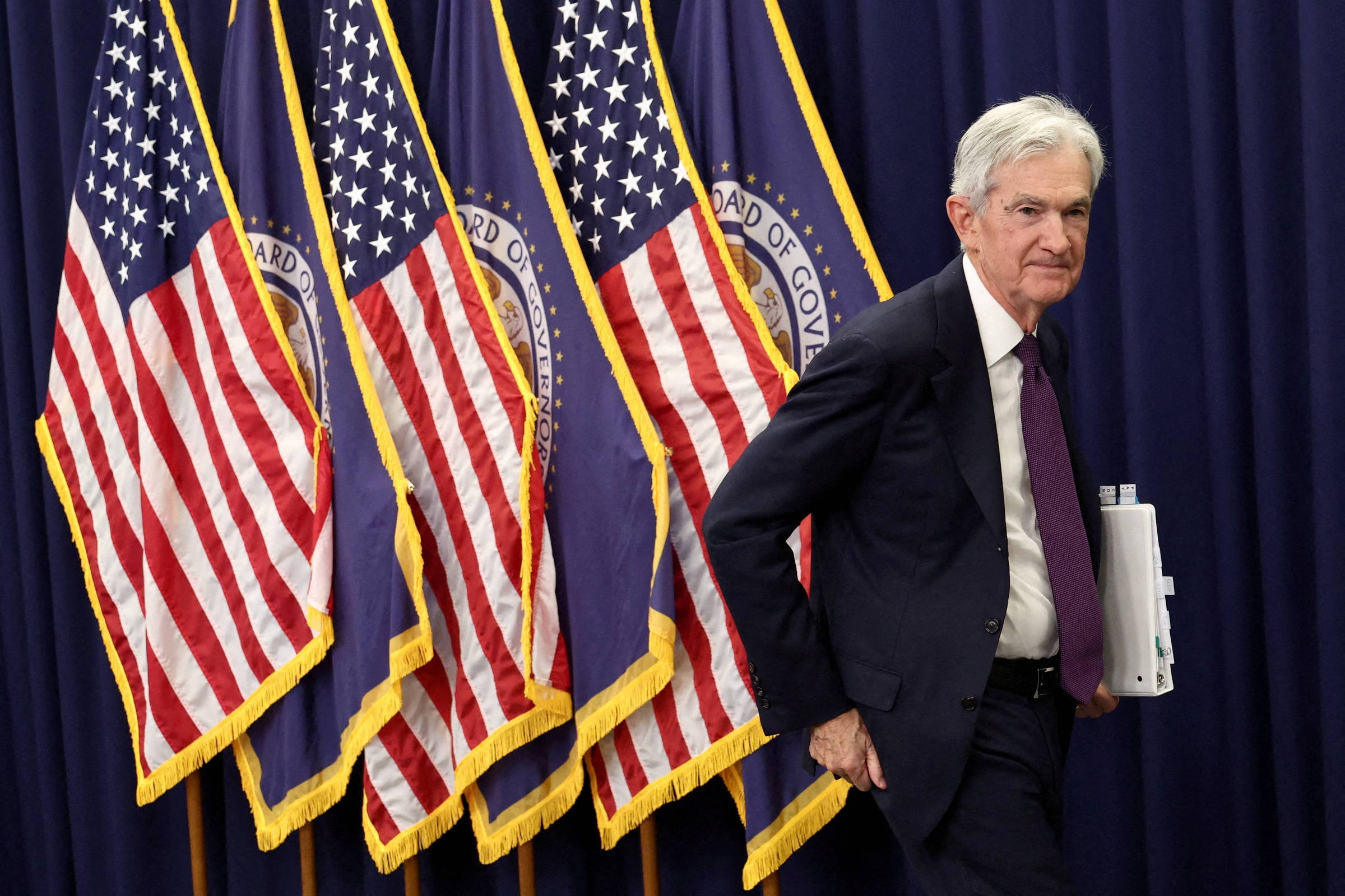 Homem de terno escuro e gravata roxa caminha à direita, segurando documentos enrolados, com cinco bandeiras americanas e do Federal Reserve ao fundo em cortina azul.