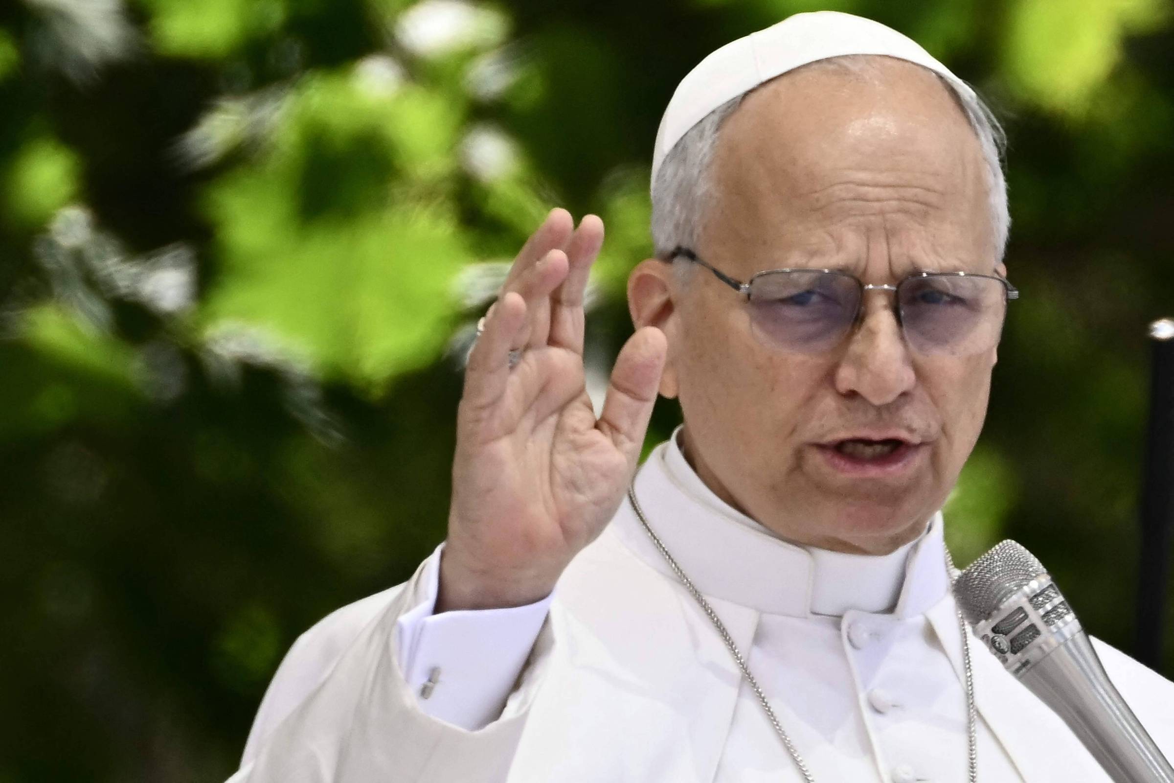 Homem idoso com vestes brancas e solidéu branco, usando óculos, fala ao microfone com a mão direita levantada. Fundo desfocado com folhagem verde.