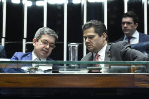 Dois homens de terno sentados lado a lado em mesa de vidro conversam atentamente. Um deles usa óculos e ambos estão focados em documentos à frente. Ao fundo, outro homem observa a cena em ambiente institucional com iluminação forte.