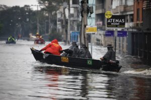 Três pessoas em barco de resgate navegam por rua inundada. Edifícios e placas comerciais visíveis ao fundo em dia nublado.