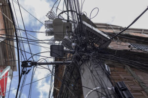 Poste de concreto com grande quantidade de fios elétricos e de telecomunicações entrelaçados e desorganizados, conectados a equipamentos e caixas instaladas no poste. Edifícios residenciais ao redor e céu parcialmente nublado ao fundo.