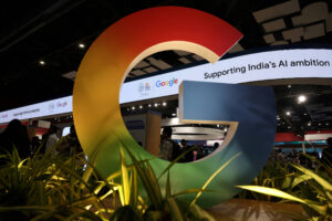 Logo tridimensional do Google em cores degradê vermelho, amarelo, verde e azul, em destaque em evento interno com iluminação artificial e plantas ao redor. Faixa digital ao fundo exibe texto