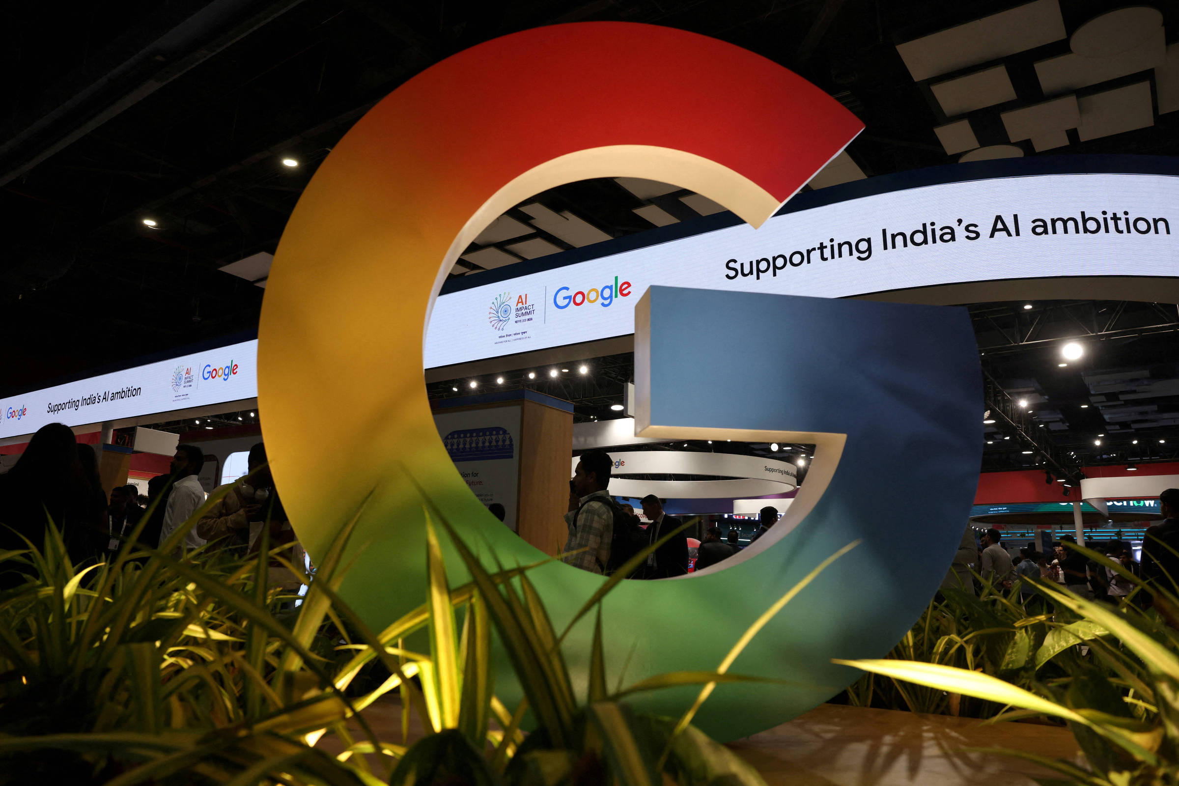 Logo tridimensional do Google em cores degradê vermelho, amarelo, verde e azul, em destaque em evento interno com iluminação artificial e plantas ao redor. Faixa digital ao fundo exibe texto