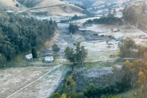 Vista aérea de área rural com duas casas próximas a uma estrada de terra, rodeadas por campos abertos e áreas de vegetação densa. Colinas suaves ao fundo e árvores dispersas completam a paisagem.