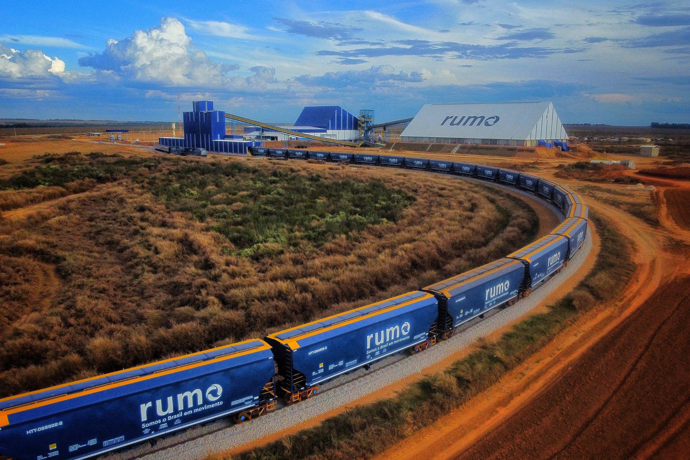 Trem de carga com vagões azuis da Rumo percorre curva em trilhos no interior, próximo a instalações industriais e vegetação seca.