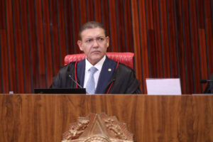 Homem sentado em cadeira vermelha atrás de bancada de madeira com brasão. Ele veste toga preta com detalhes vermelhos e gravata azul, em ambiente com painéis de madeira ao fundo.