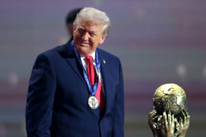 Ex-presidente dos EUA veste terno azul e gravata vermelha, usando medalha prateada no pescoço, sorrindo ao lado de troféu dourado com mãos esculpidas.