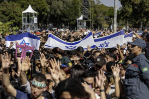 A imagem mostra uma grande multidão reunida em um evento ao ar livre, com várias pessoas segurando bandeiras de Israel. Algumas pessoas estão com as mãos levantadas, enquanto outras estão visíveis em primeiro plano. Ao fundo, há estruturas de palco e árvores. Um segurança, vestido com uniforme, observa a cena.