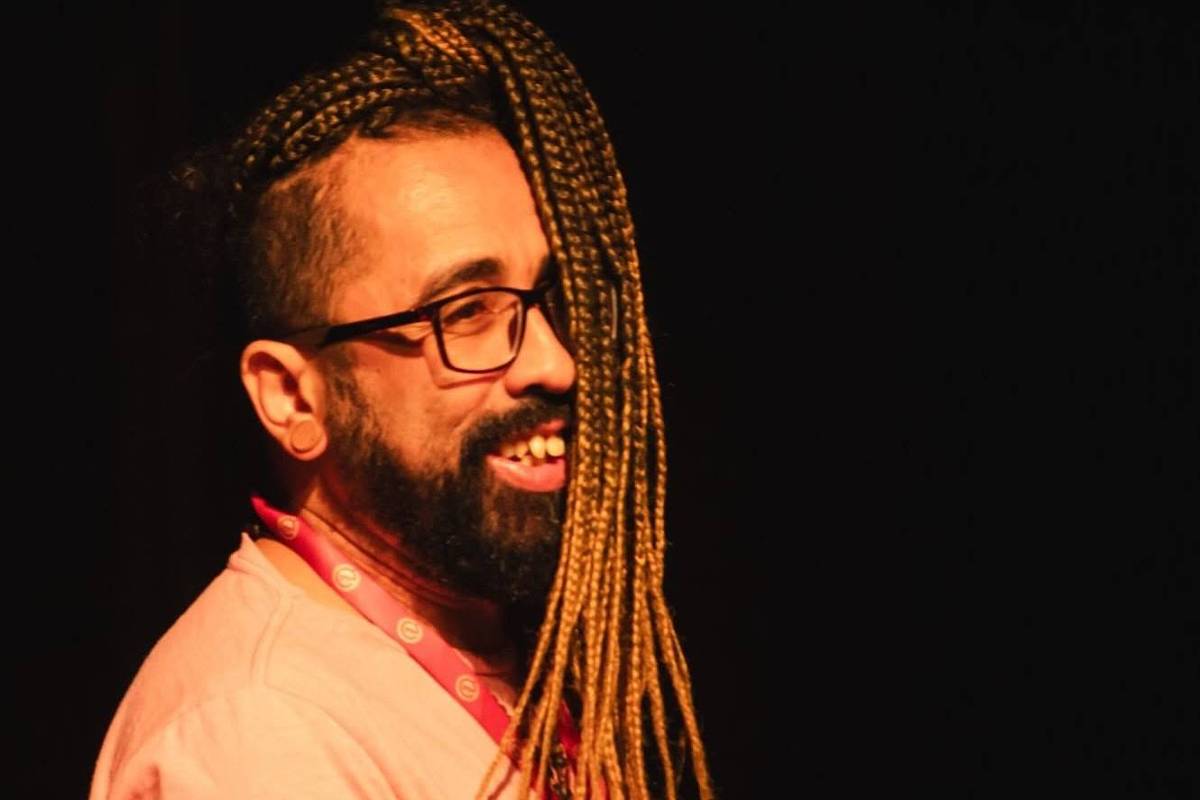 Homem com barba e óculos de armação escura sorri olhando para a esquerda. Ele usa camiseta clara e tem tranças longas e loiras que caem sobre o ombro direito, com o cabelo raspado nas laterais. Fundo preto destaca o rosto e o cabelo.