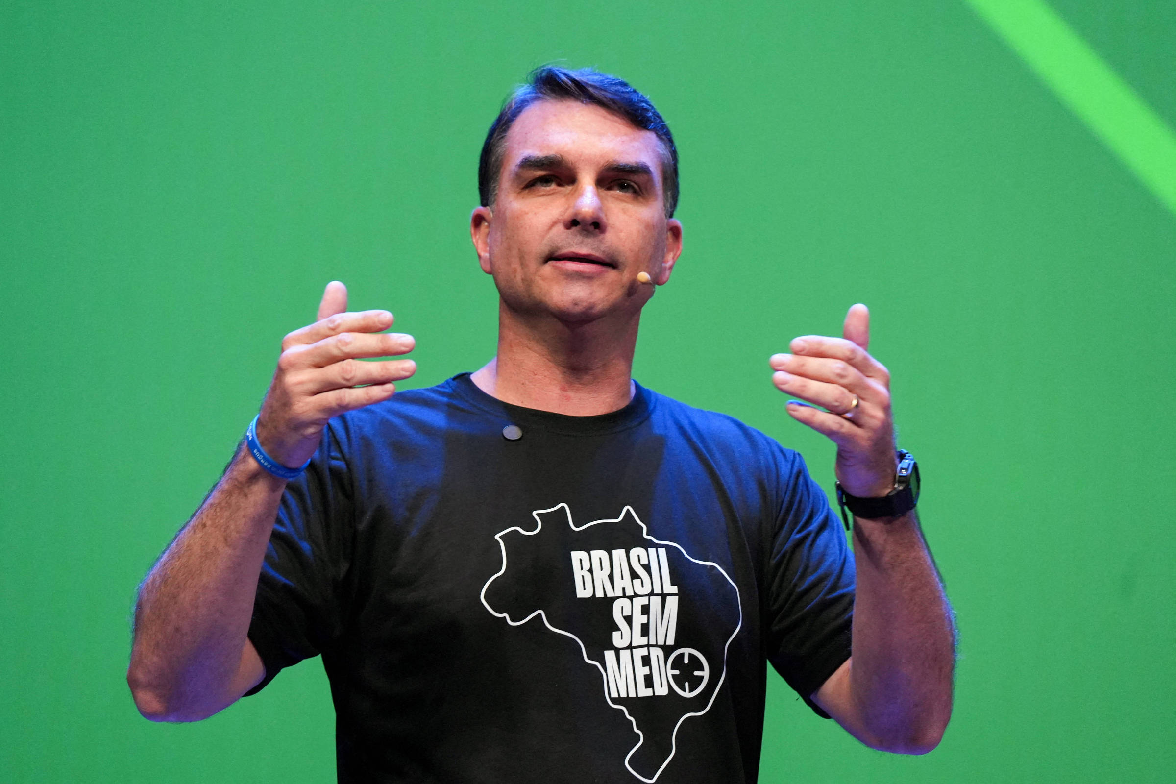 Homem de meia-idade com microfone de lapela gesticula com as mãos abertas em frente a fundo verde. Ele veste camiseta preta com mapa do Brasil e texto