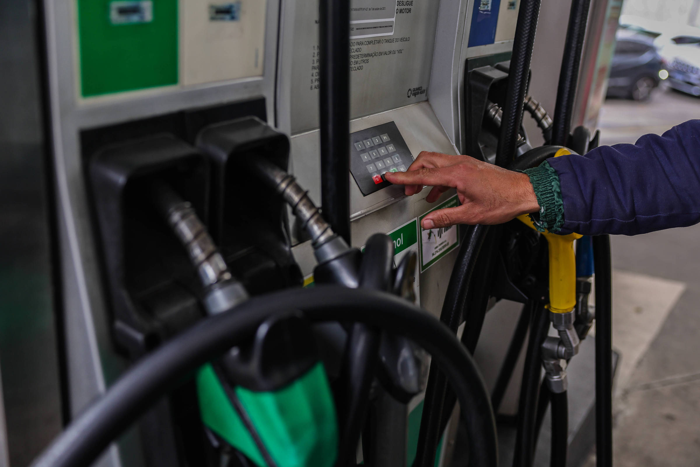 A imagem mostra uma bomba de combustível em um posto de gasolina. Uma mão está pressionando um botão na bomba, enquanto as mangueiras de abastecimento estão visíveis, com uma delas destacada em verde e outra em amarelo. Ao fundo, é possível ver um carro estacionado.