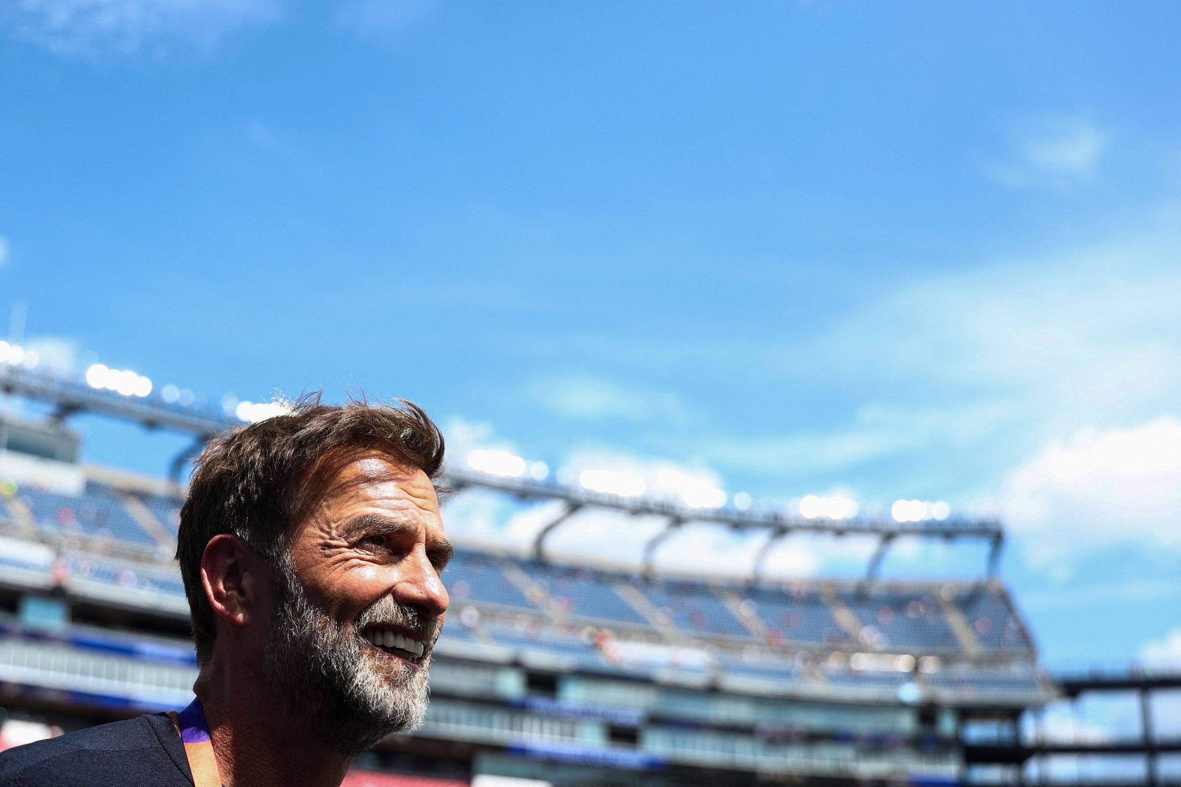 Homem de barba e cabelo castanho claro sorri olhando para a direita, com arquibancadas de estádio ao fundo sob céu azul com poucas nuvens.