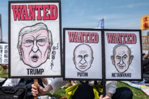 Três cartazes com caricaturas de Donald Trump, Vladimir Putin e Benjamin Netanyahu exibidos em protesto ao ar livre. Cada cartaz tem a palavra