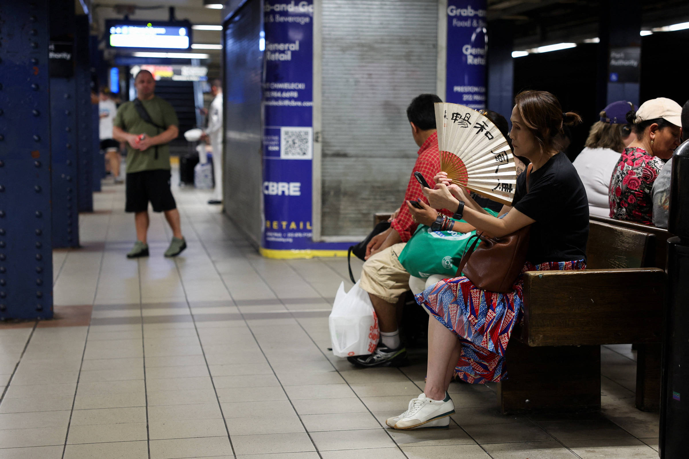 Cinco pessoas sentadas em banco de madeira em estação de metrô. Mulher à direita usa leque com caracteres asiáticos. Homem ao fundo caminha pelo corredor da plataforma.