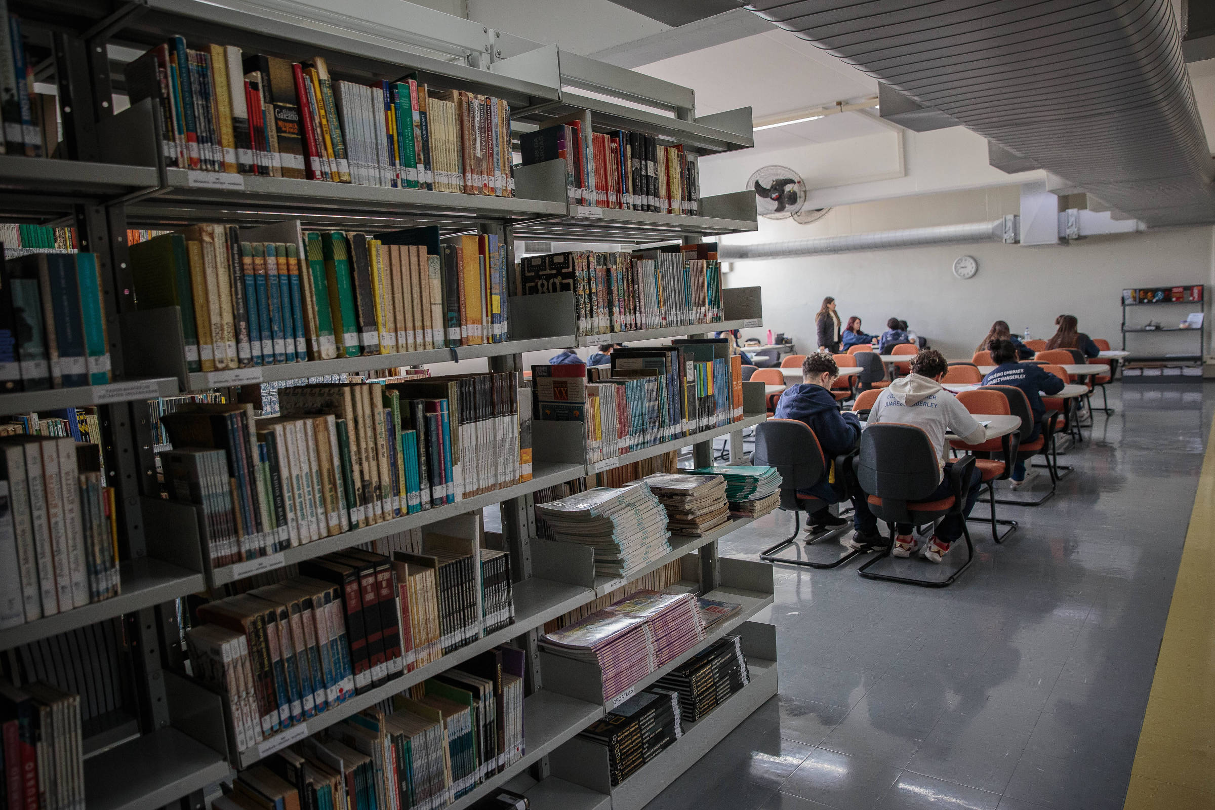 A imagem mostra o interior de uma biblioteca. À esquerda, há estantes de livros organizados, com capas visíveis. À direita, um grupo de estudantes está sentado em mesas, alguns com laptops, em um ambiente de estudo. Ao fundo, há um relógio na parede e uma janela que permite a entrada de luz natural. O piso é de cerâmica e a iluminação é artificial.