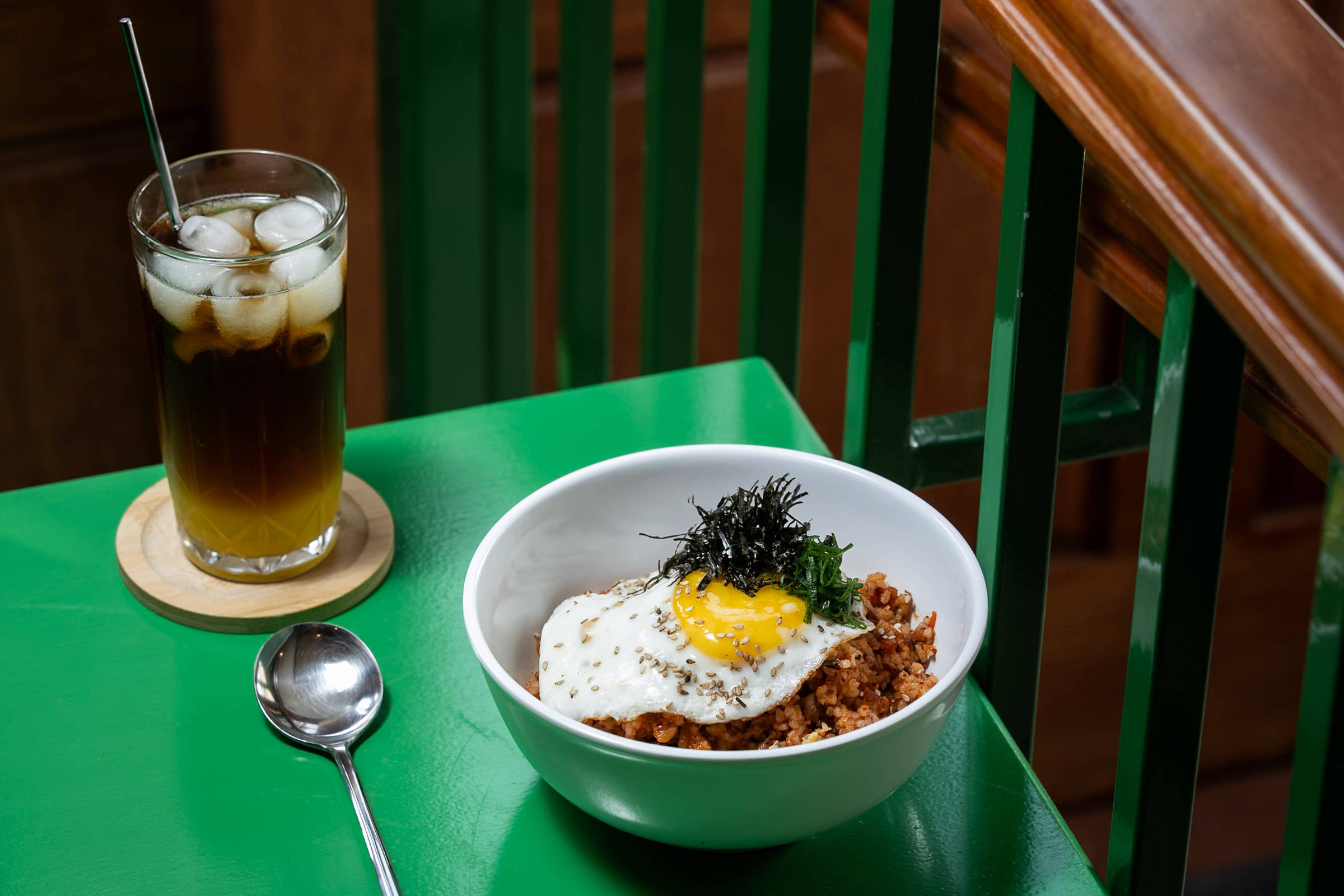 Tigela branca com arroz temperado coberto por ovo frito com gema mole e algas secas, sobre mesa verde. Ao lado, copo alto com chá gelado e gelo, com colher ao lado.