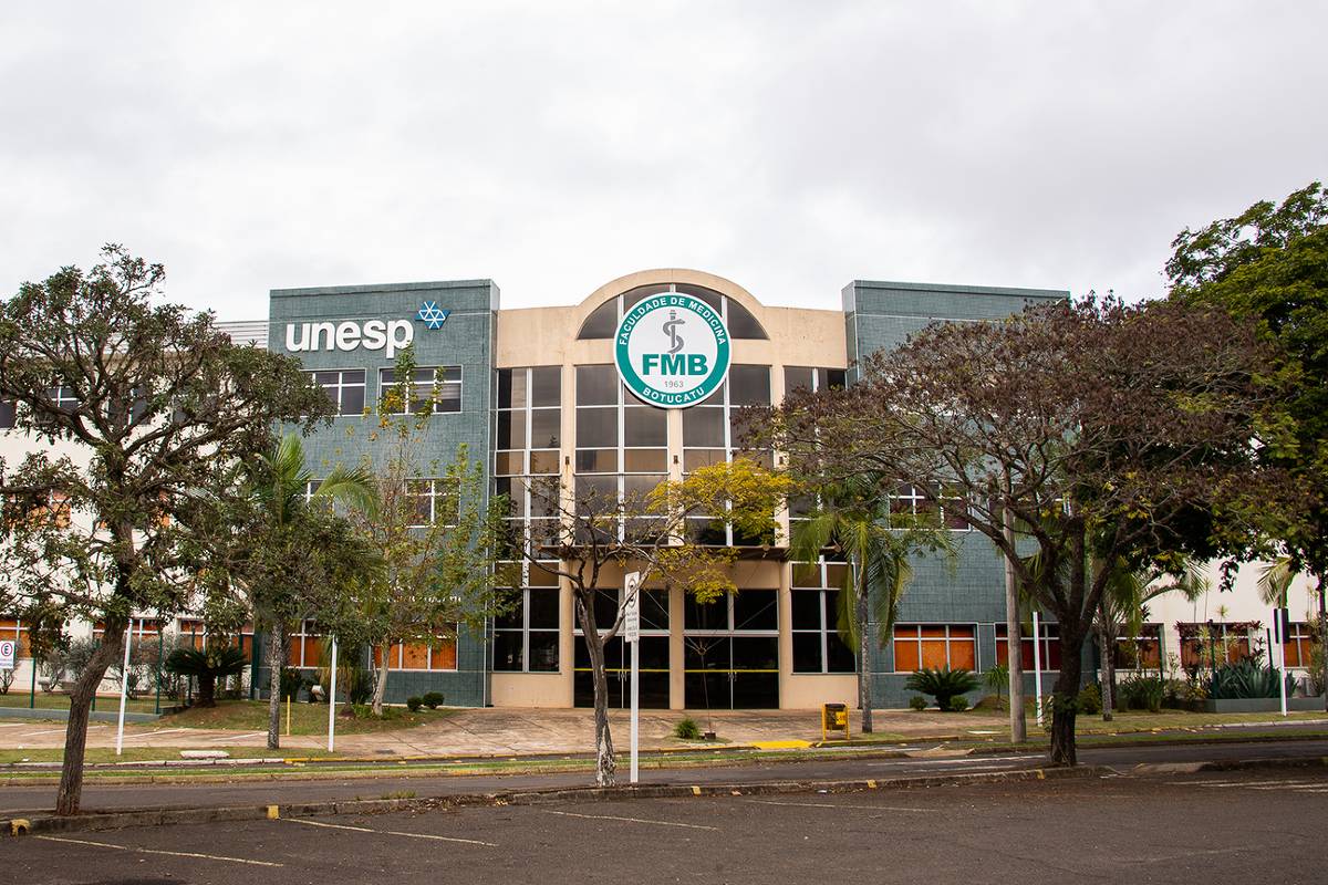 A imagem mostra a fachada de um edifício da UNESP, com o logotipo da universidade visível. O prédio é de dois andares, com janelas amplas e uma entrada central. À frente, há árvores e um espaço pavimentado. O céu está nublado.