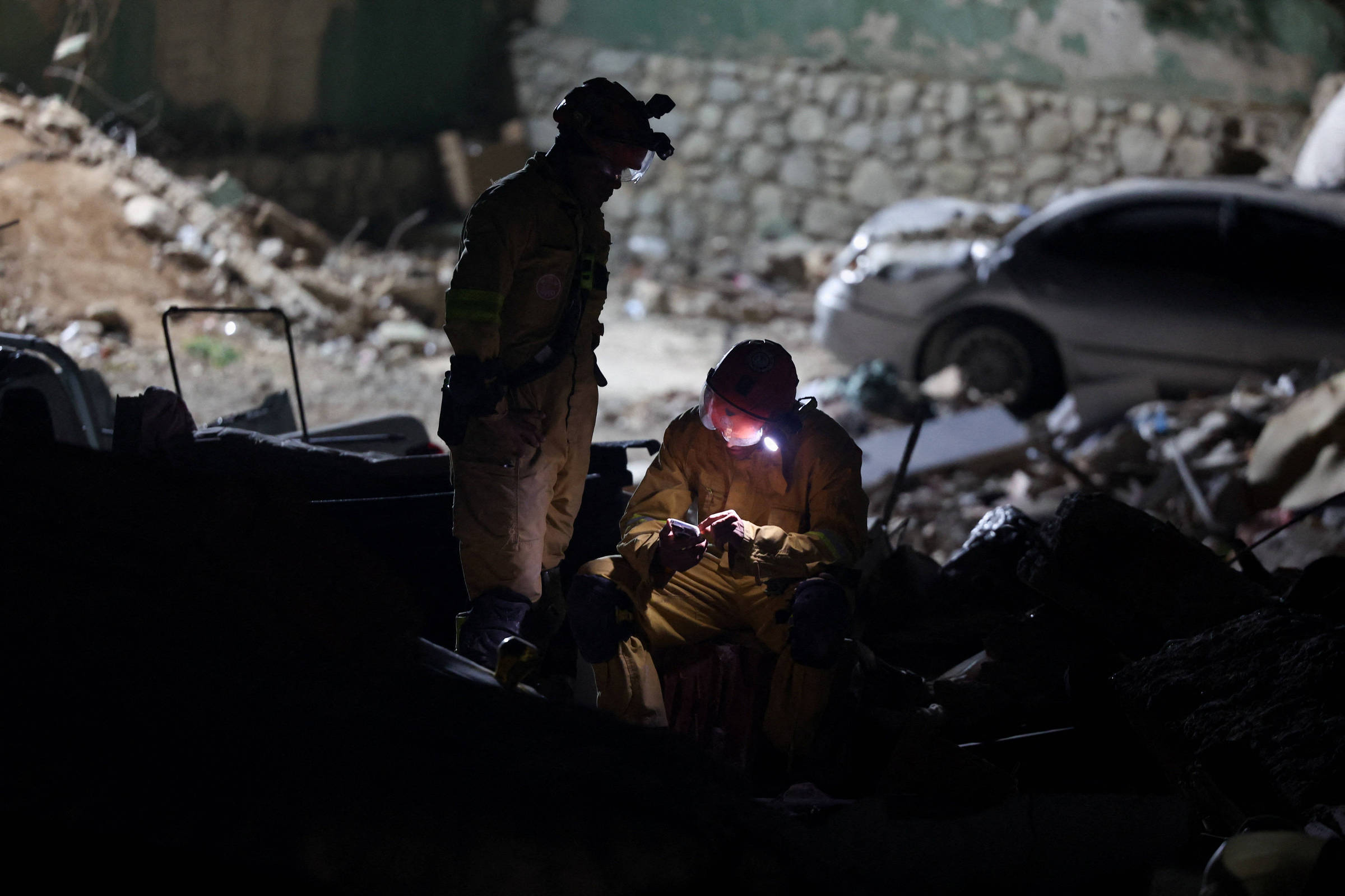 Dois socorristas em roupas de proteção e capacetes com lanternas trabalham entre destroços à noite. Um está agachado examinando algo com luz direta, enquanto o outro observa em pé. Ao fundo, carro danificado e parede de pedras.