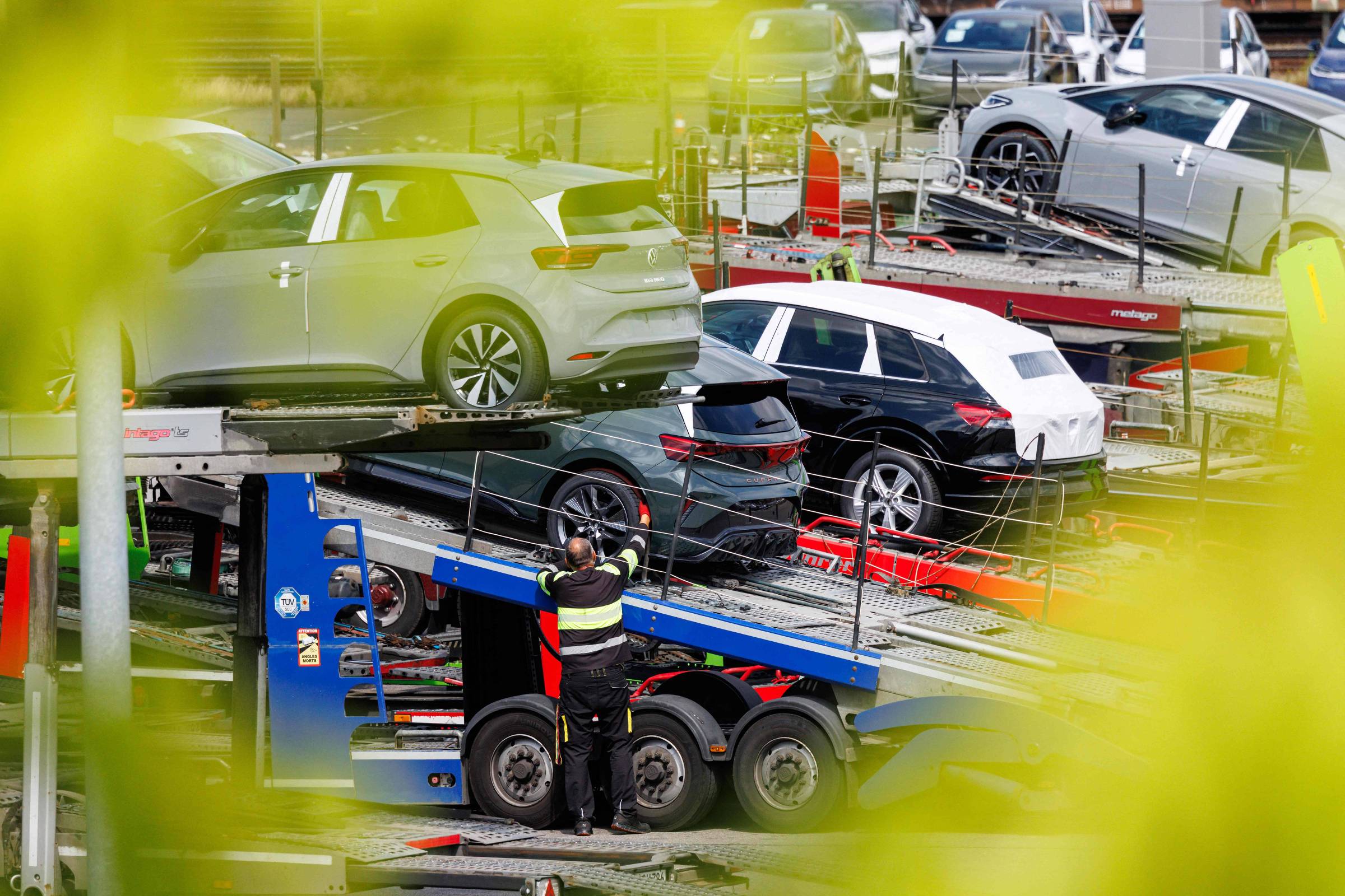 Três carros elétricos estacionados em plataforma de caminhão transportador em área externa. Homem com colete refletivo ajusta equipamento na lateral do caminhão. Veículos são de cores prata, preta e cinza, com ambiente urbano ao fundo.