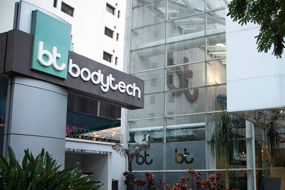 Fachada da academia Bodytech com logo verde e branco em painel escuro. Prédio de vidro e metal com plantas ao redor e iluminação interna visível.