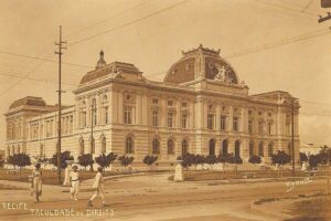 Edifício clássico da Faculdade de Direito em Recife com arquitetura neoclássica, janelas altas e detalhes ornamentais. Três pessoas caminham na calçada em frente, árvores pequenas alinham a rua e postes de eletricidade com fios cruzam o céu.