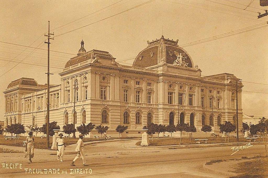 Edifício clássico da Faculdade de Direito em Recife com arquitetura neoclássica, janelas altas e detalhes ornamentais. Três pessoas caminham na calçada em frente, árvores pequenas alinham a rua e postes de eletricidade com fios cruzam o céu.