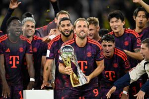 Jogadores do Bayern de Munique vestindo uniforme roxo com detalhes vermelhos posam juntos em comemoração. Um jogador no centro segura um troféu dourado com esfera prateada no topo. Confetes dourados caem ao redor, e todos exibem expressões de alegria.