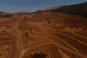 Vista aérea de mineração a céu aberto com terra vermelha escavada em níveis. Caminho de terra percorre a área, com vegetação esparsa nas bordas e colinas ao fundo.
