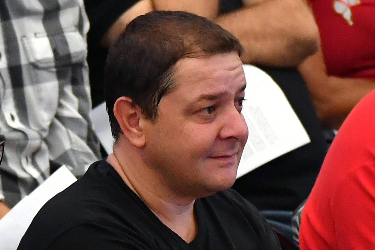 Homem de cabelo curto e escuro veste camiseta preta e está sentado, olhando para a direita. Ao fundo, outras pessoas aparecem parcialmente, uma com camisa xadrez preta e branca e outra com camiseta vermelha.
