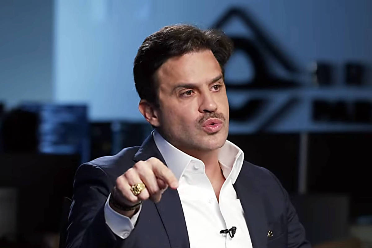 Homem de cabelo escuro e bigode, vestido com camisa branca e blazer azul escuro, gesticula com a mão direita enquanto fala em ambiente interno com iluminação suave e fundo desfocado.