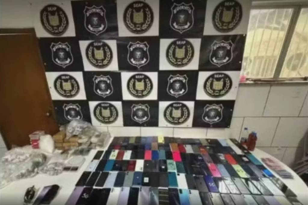 Mesa com dezenas de celulares organizados em fileiras, além de pacotes e objetos diversos. Ao fundo, parede com bandeira preta e branca exibindo emblemas da polícia.