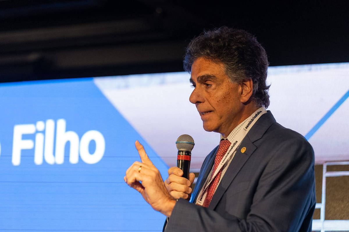 Homem de terno azul e gravata vermelha segura microfone e gesticula com a mão direita durante apresentação. Fundo azul com texto parcialmente visível