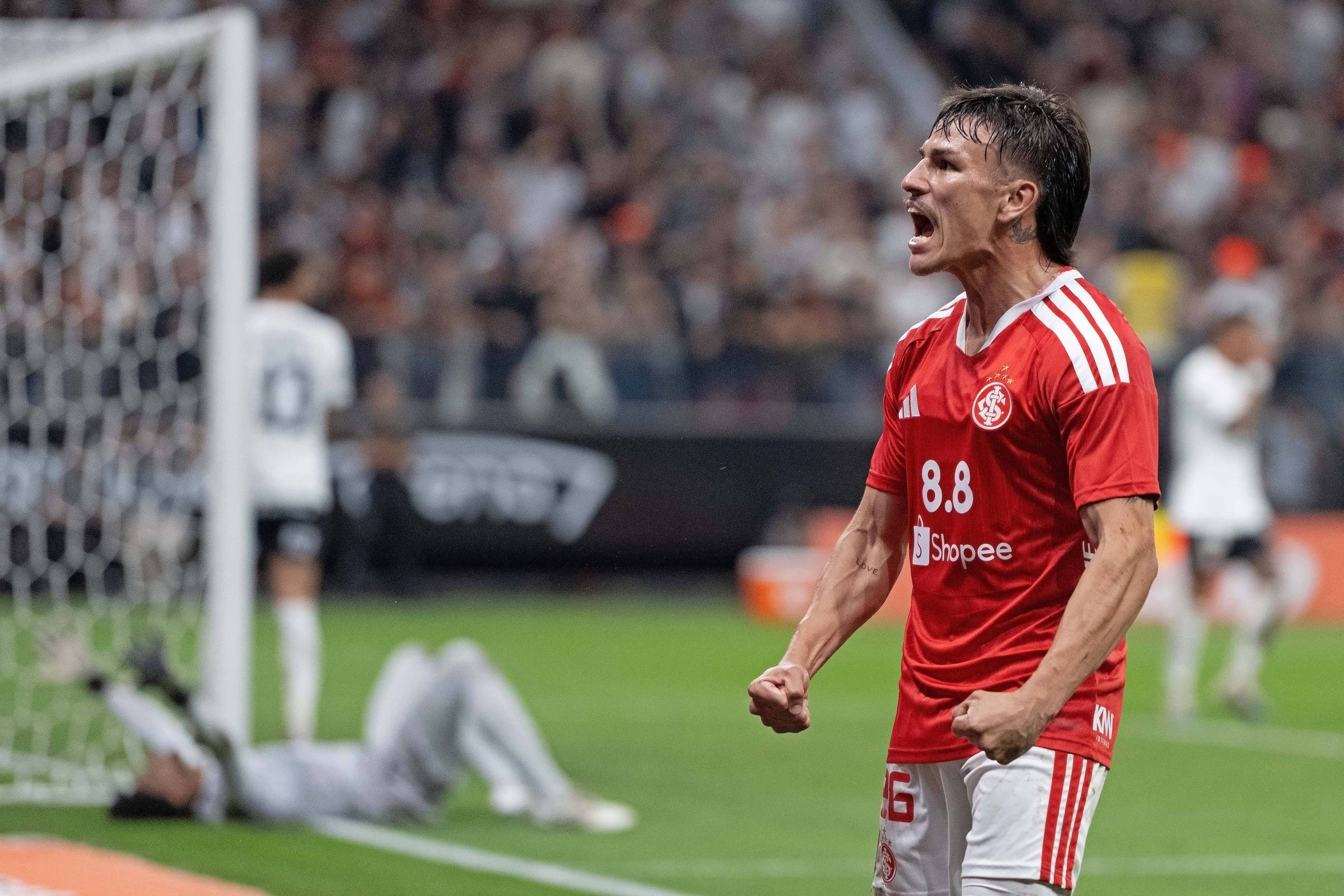 Jogador com uniforme vermelho e branco comemora com expressão intensa próximo à área do gol. Goleiro adversário está caído no chão perto da trave, enquanto outros jogadores e torcida aparecem desfocados ao fundo.