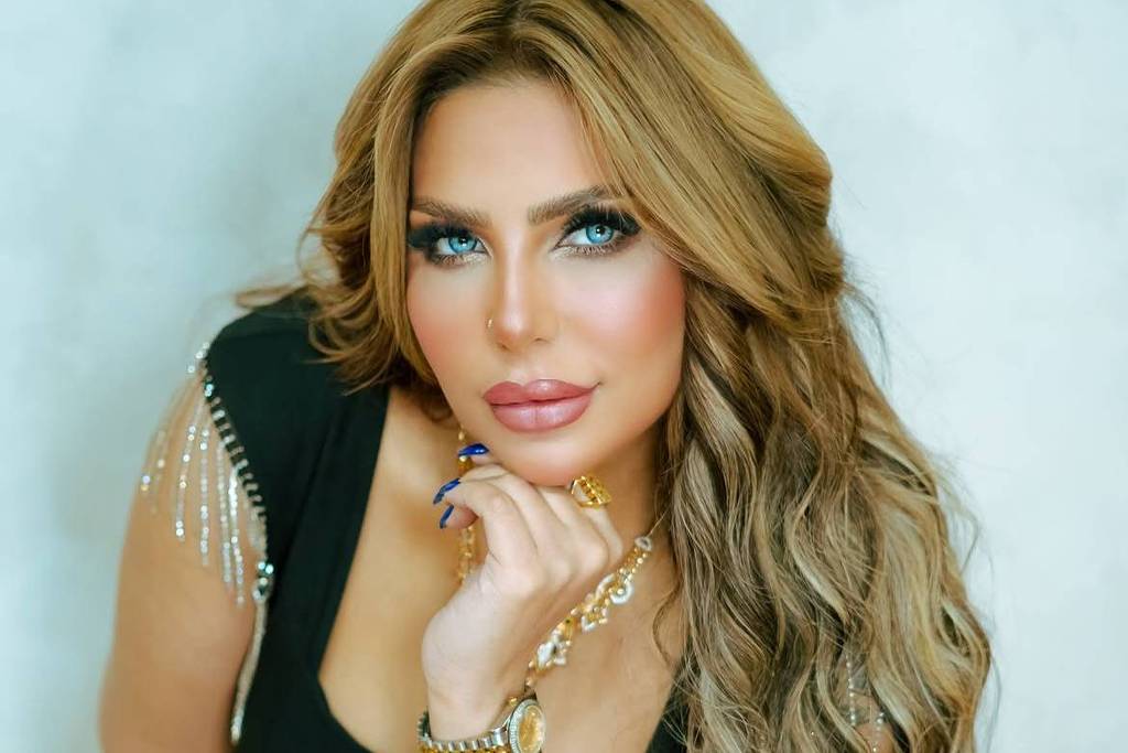 Mulher loira de cabelos longos e ondulados usa vestido preto com detalhes de franjas prateadas nos ombros. Ela está apoiando o queixo na mão direita, exibindo pulseiras douradas e anel, com fundo neutro claro.