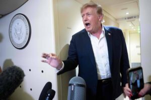Donald Trump, ex-presidente dos EUA, fala com expressão enfática em corredor interno. Ele veste terno azul escuro e camisa branca sem gravata. Microfones e câmeras estão à sua frente, e o selo presidencial está visível na parede ao lado.