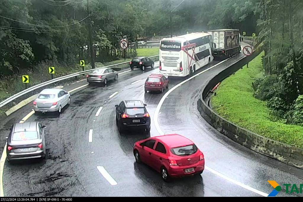 A imagem mostra uma estrada em curva com tráfego de veículos durante uma chuva. Um ônibus de dois andares está na pista, enquanto vários carros, incluindo um carro vermelho, estão se movendo. A vegetação ao redor é densa e verde, e a pista parece molhada devido à chuva.