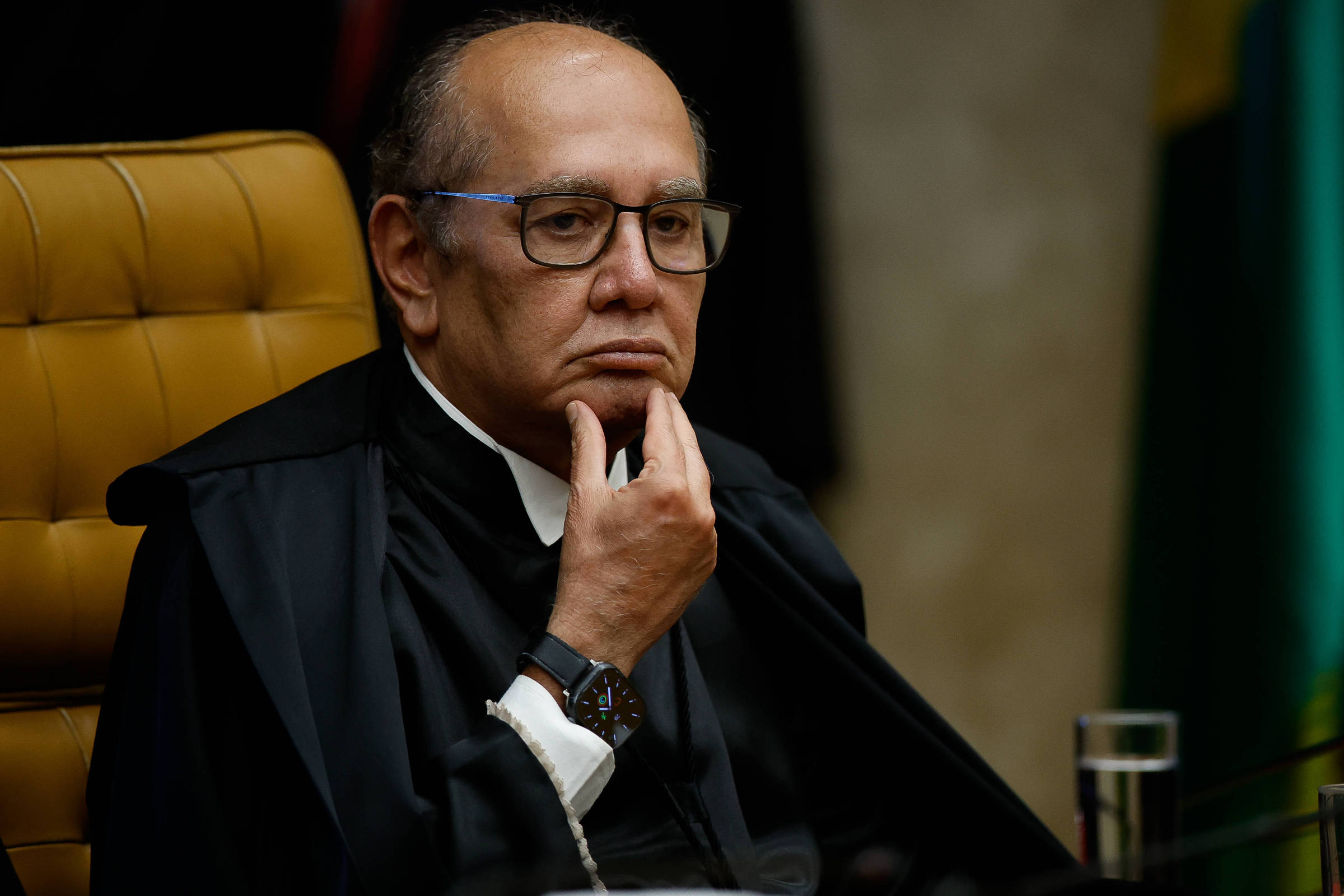 Ministro do Supremo Tribunal Federal sentado em cadeira de couro marrom, vestindo toga preta e óculos, com expressão pensativa e mão no queixo.