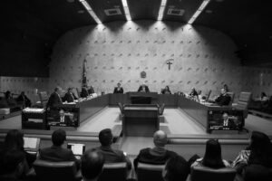 Plenário do Supremo Tribunal Federal com ministros sentados em semicírculo ao redor de mesa central. Público observa a sessão em cadeiras na parte inferior da imagem. Paredes com textura e símbolos oficiais ao fundo.