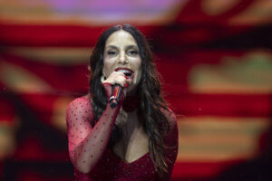 Cantora de cabelos longos e ondulados usa vestido vermelho com mangas transparentes e detalhes em poás, segurando microfone vermelho enquanto canta em palco iluminado.