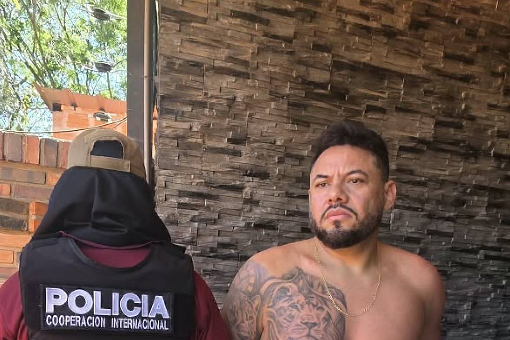 Homem sem camisa e de bermuda branca com estampa preta está algemado de costas para a parede de pedra. Policial com colete à prova de balas escrito