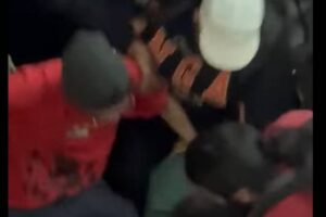 Cinco pessoas cercam e seguram um indivíduo vestido de preto no chão. Uma pessoa usa boné branco e outra veste camiseta vermelha. Ambiente interno com pouca iluminação.
