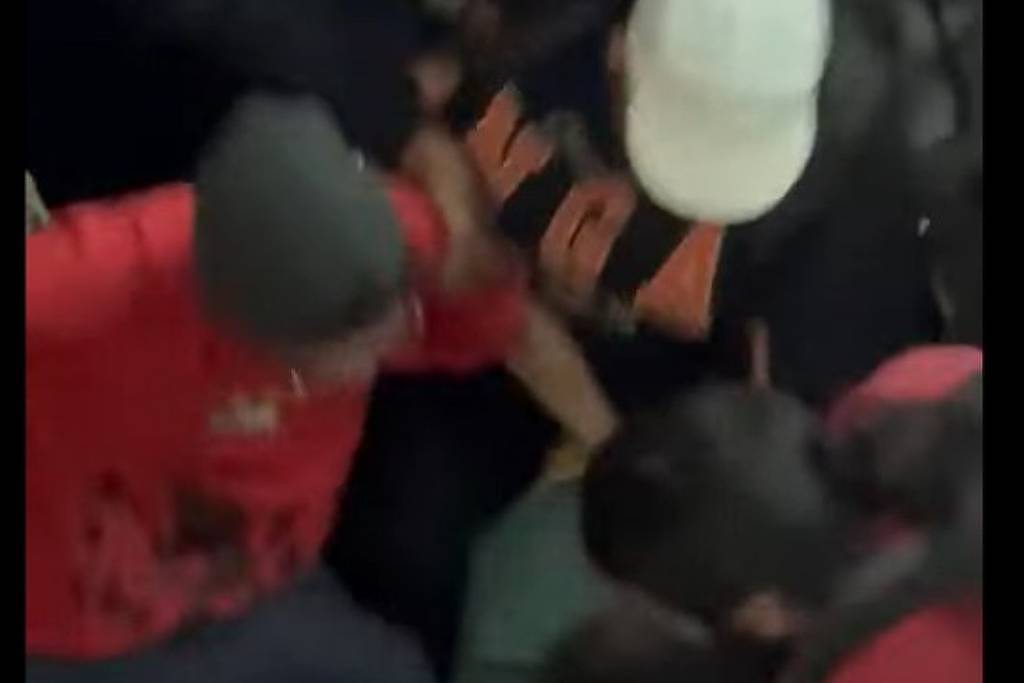 Cinco pessoas cercam e seguram um indivíduo vestido de preto no chão. Uma pessoa usa boné branco e outra veste camiseta vermelha. Ambiente interno com pouca iluminação.
