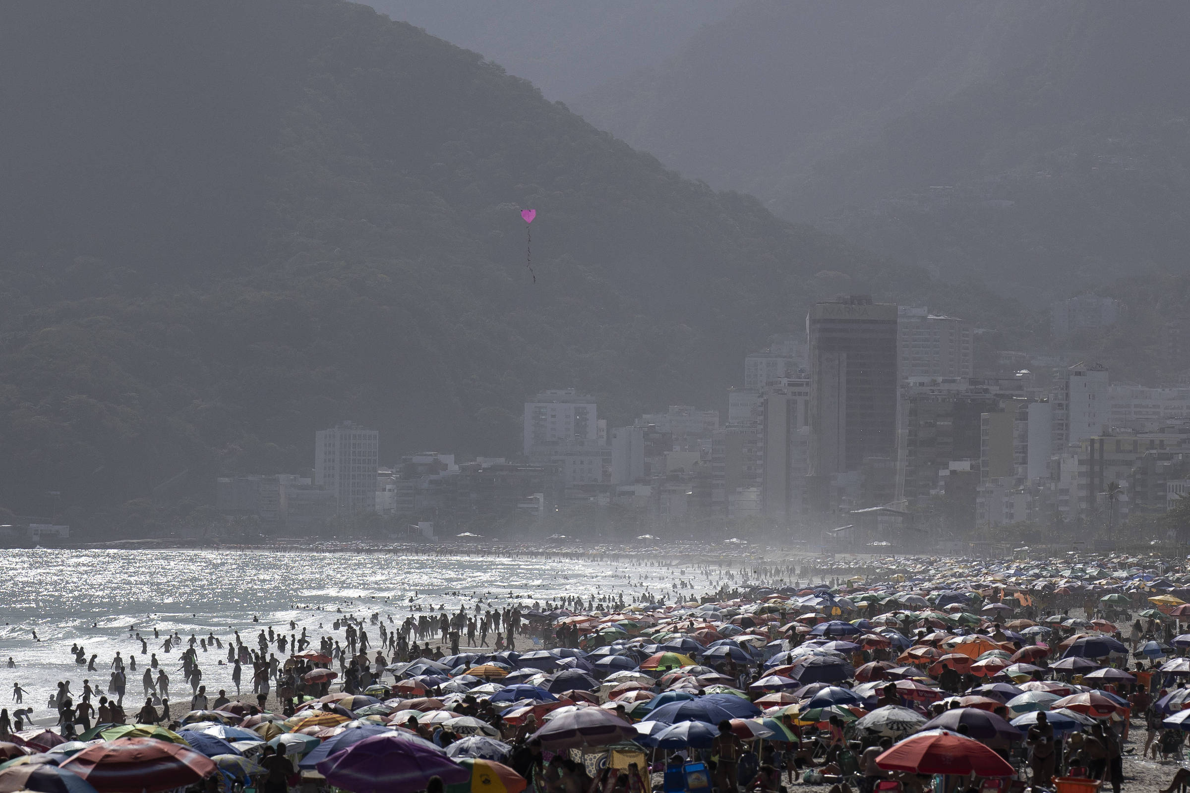 Multidão de pessoas na areia e no mar de uma praia urbana com muitos guarda-sóis coloridos. Ao fundo, prédios altos e morros cobertos por vegetação sob céu nublado.